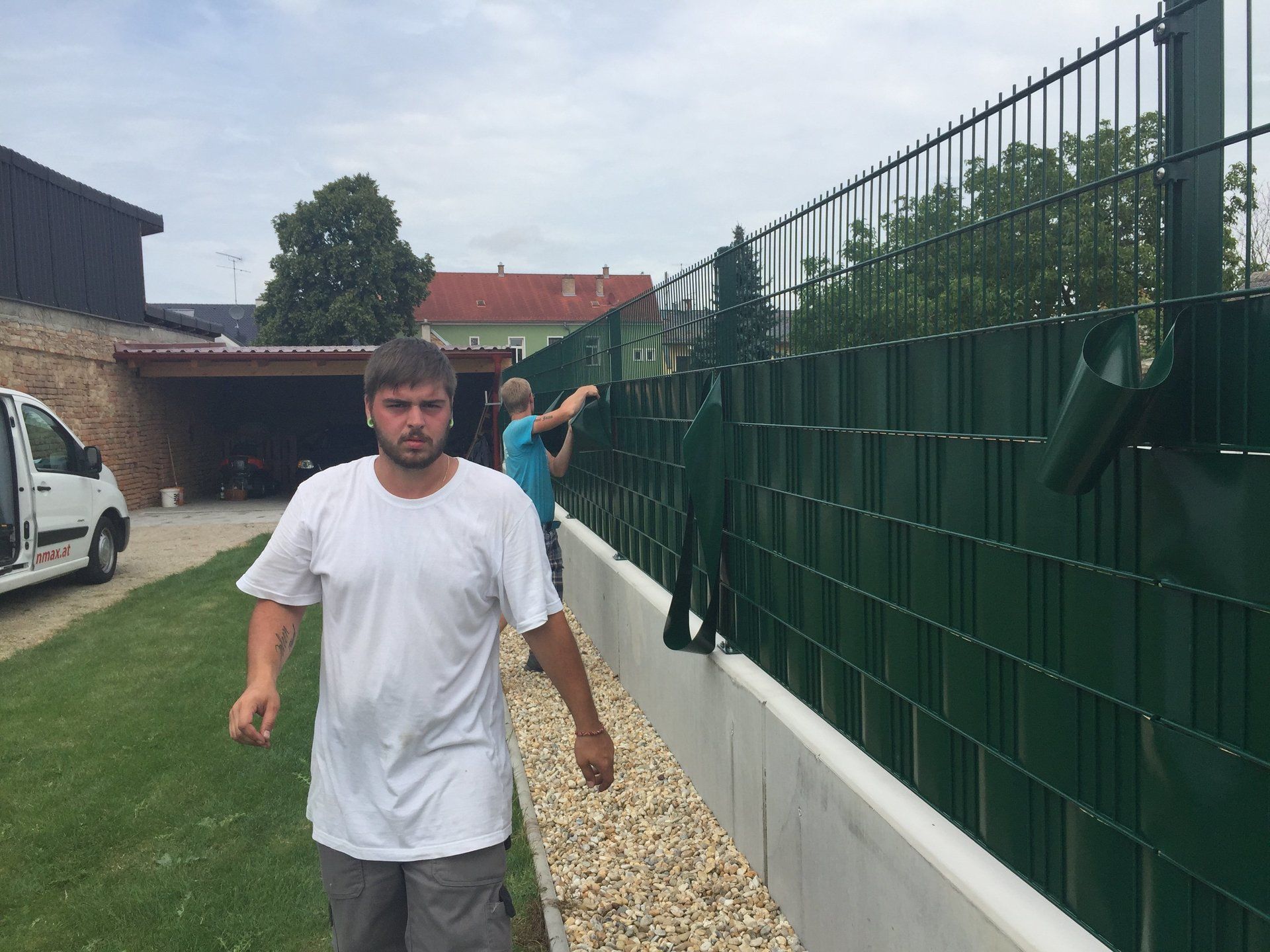 Ein Mann in einem weißen Hemd steht neben einem grünen Zaun.