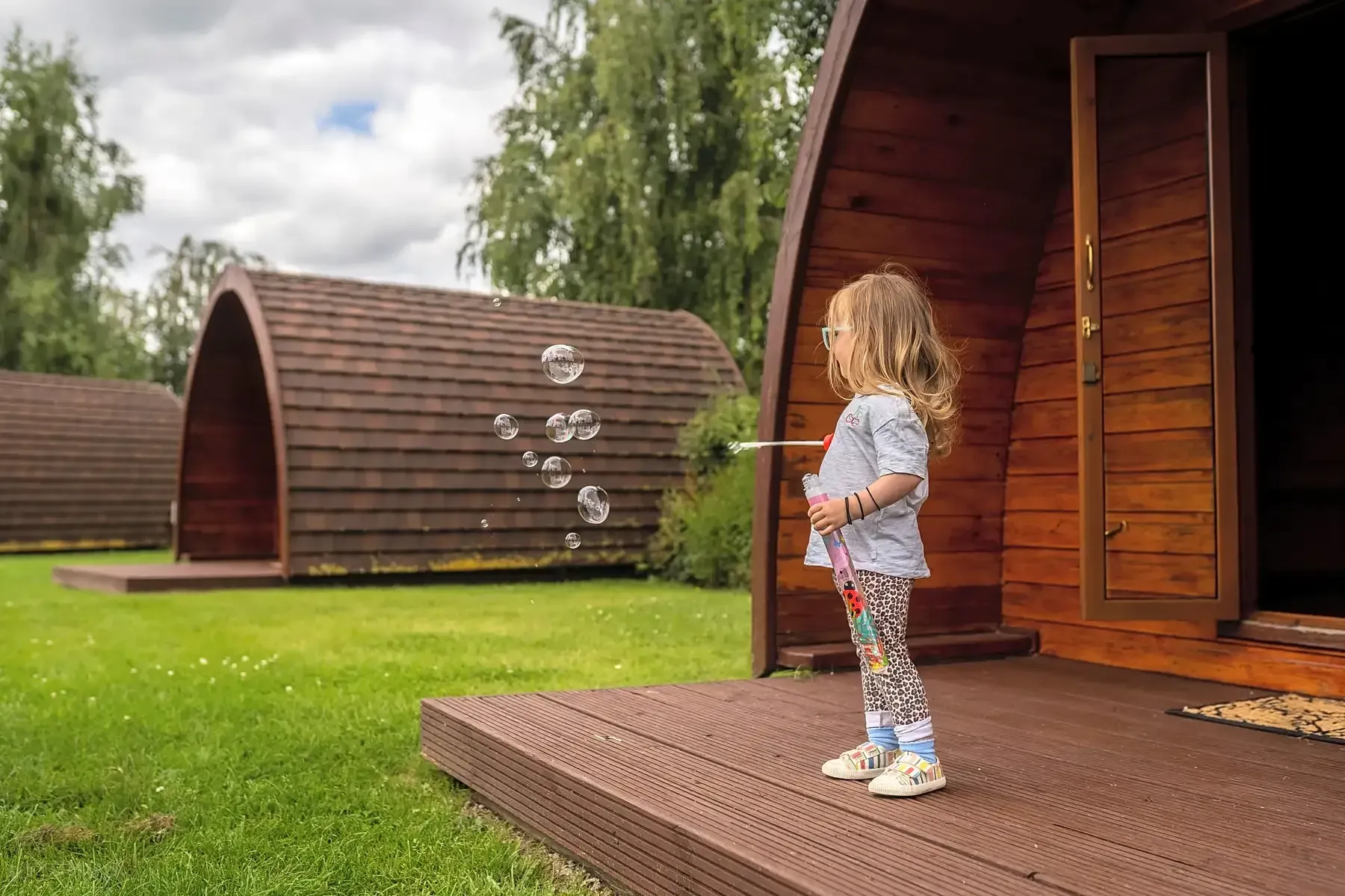 A young child blowing bubbles on a wooden deck in front of a camping pod.