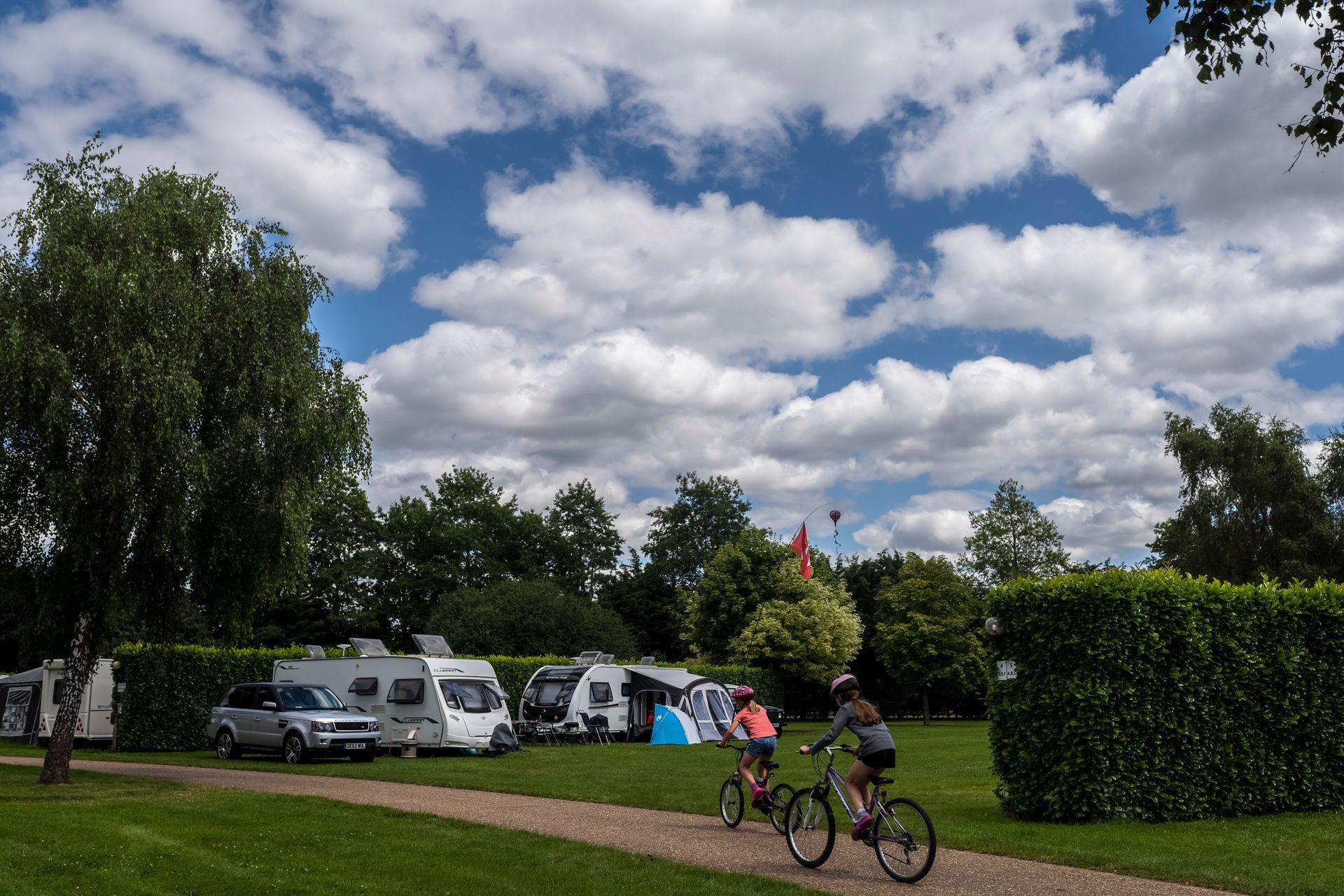 Campground scene with RVs, trees, cyclists on a path, and a cloudy blue sky.