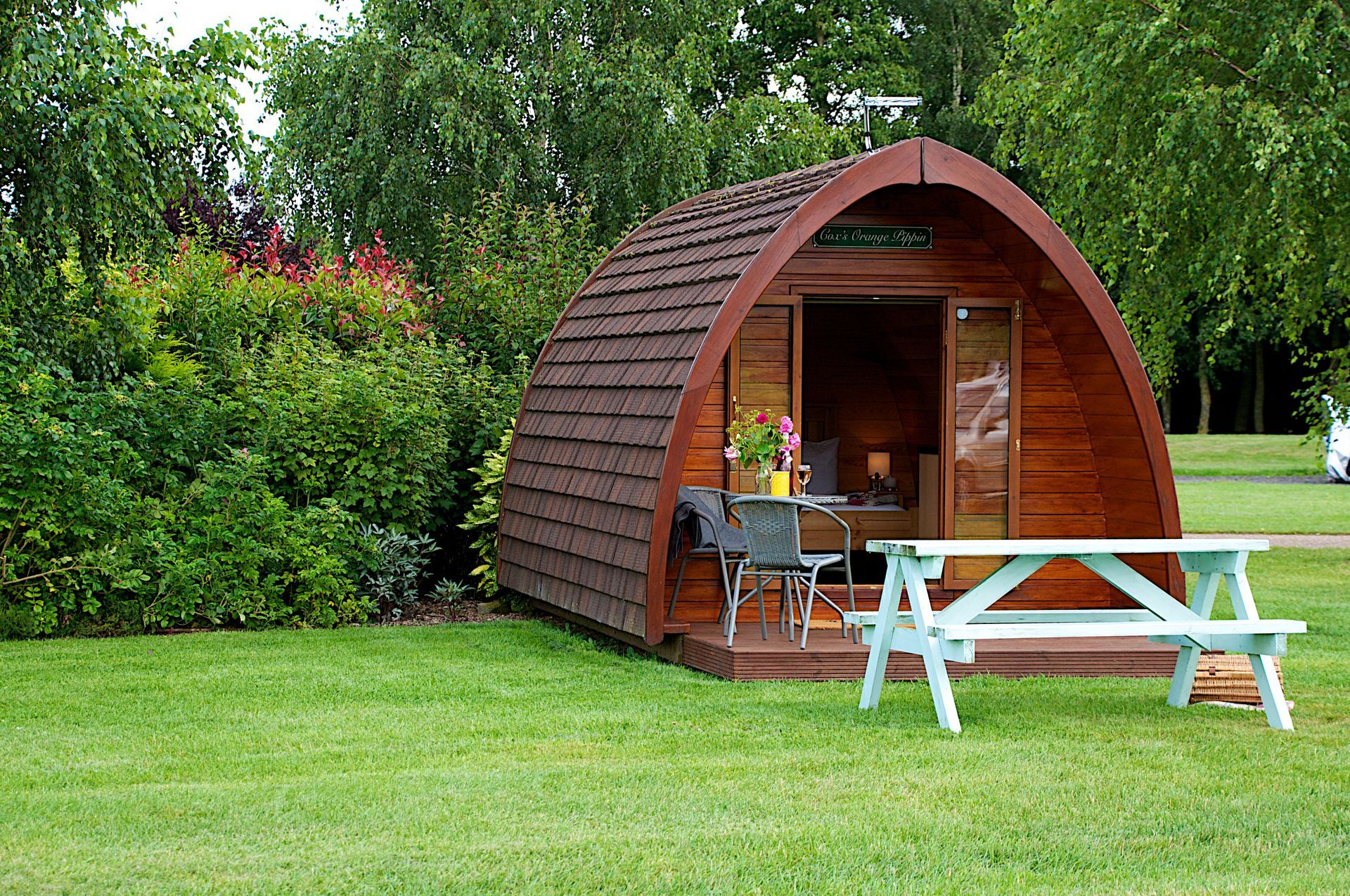 Wooden camping pod with open door, picnic table, and shrubs.