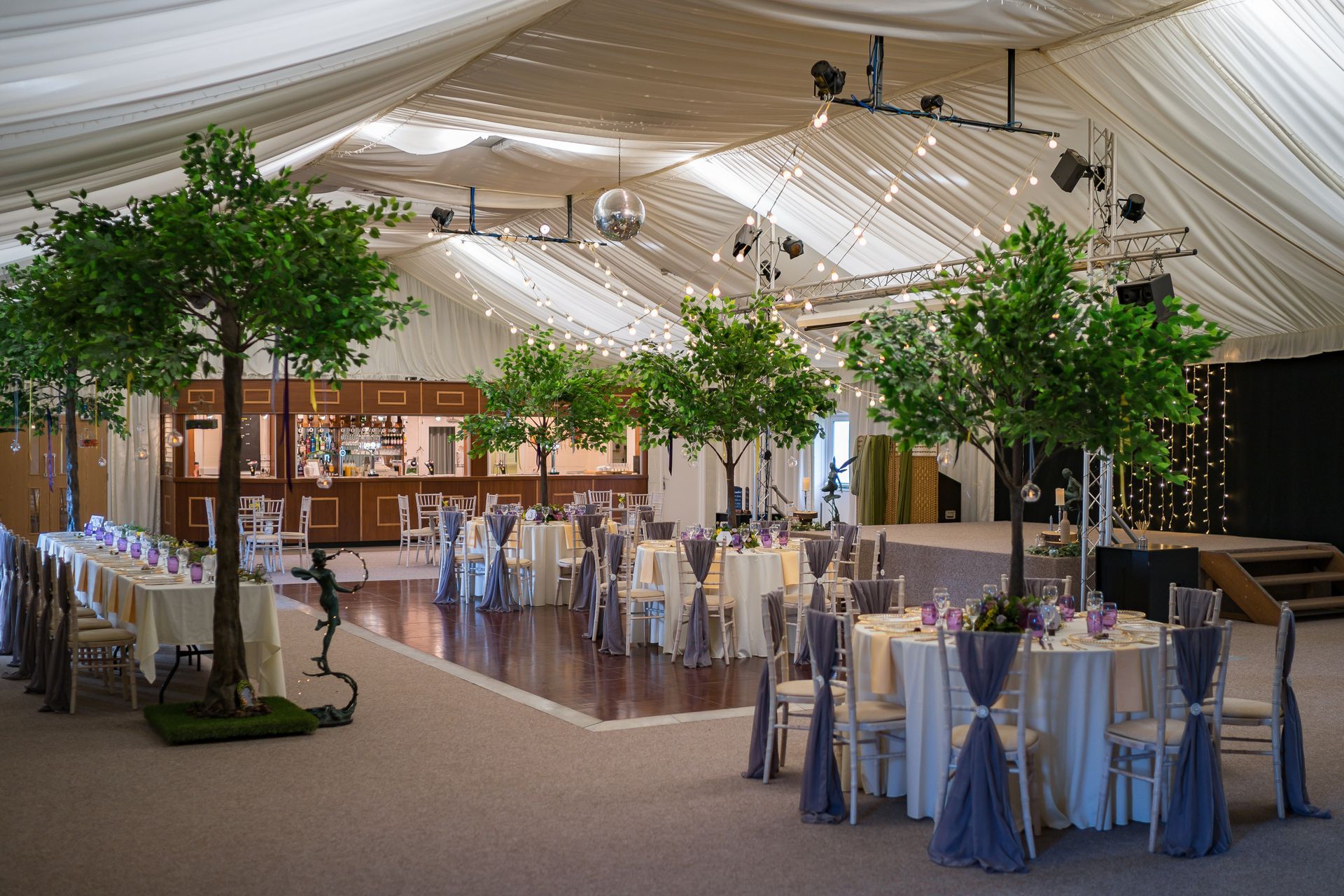 Wedding reception inside hall, with tables, trees, and a bar.  White, purple, and green colors.