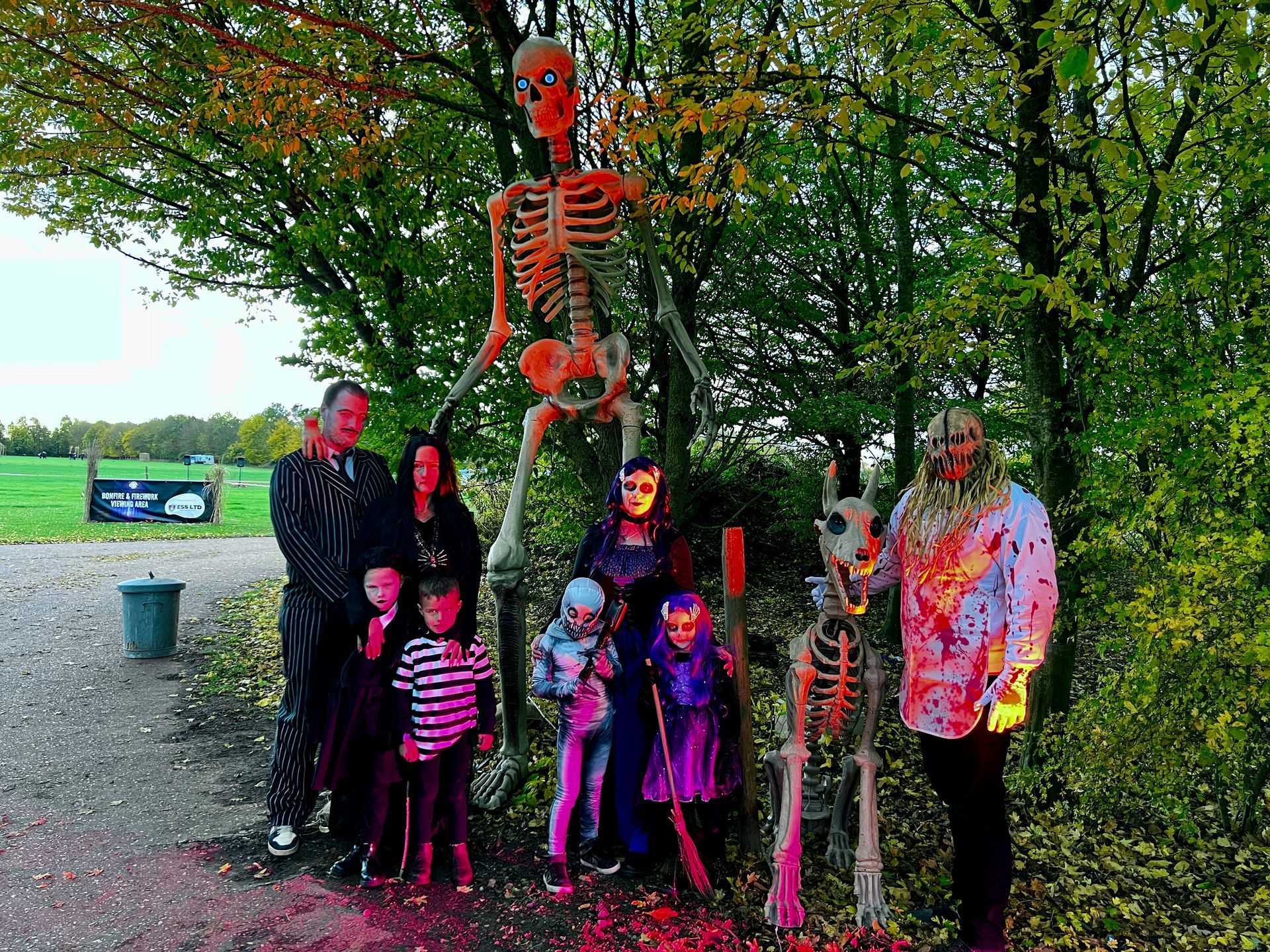 Group of costumed people pose with Halloween decorations, including a large skeleton, in front of a green field.