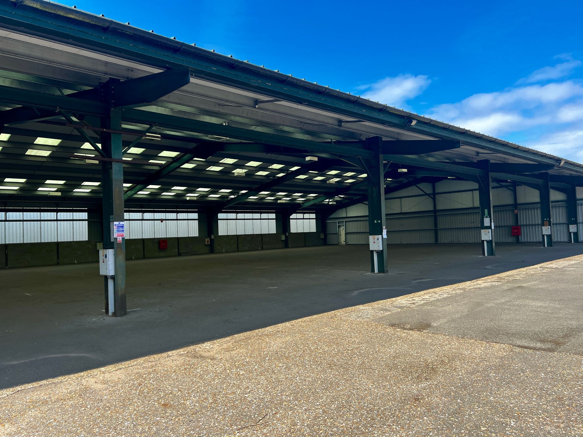 Open-air, green-framed building with a corrugated roof. Empty interior, gravel ground, and blue sky.