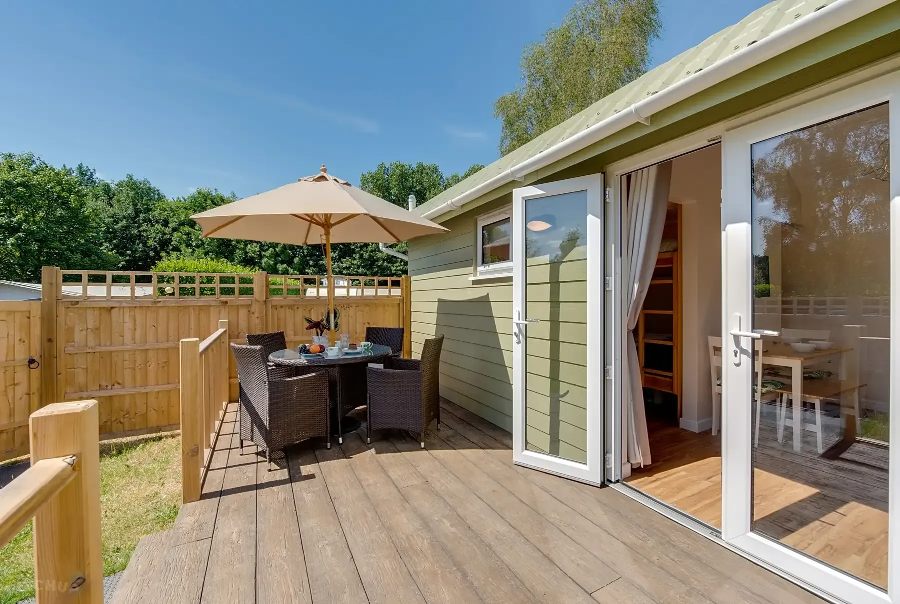 Wooden patio with outdoor dining set and open French doors to a light-green building on a sunny day.