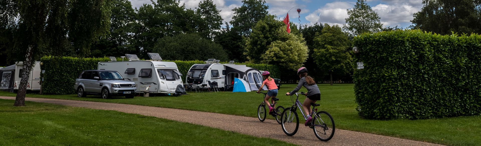 Two children cycling along a path in a lush green countryside setting with two caravans in the background