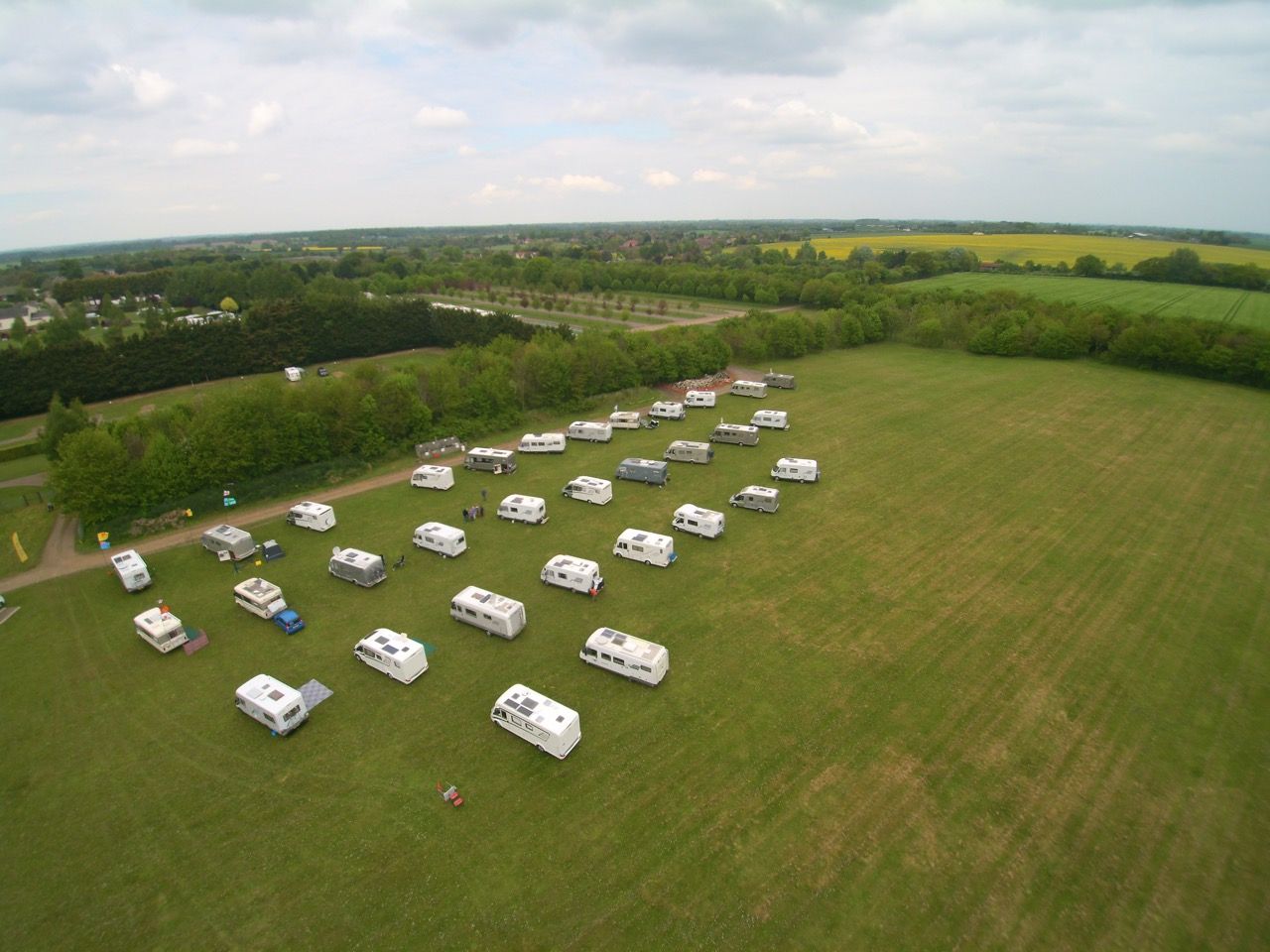 Aerial view of many white campervans parked in a grassy field. Trees and fields are in the background.