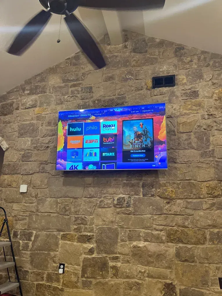Large TV mounted on a stone wall, showing a colorful streaming service menu; ceiling fan above.