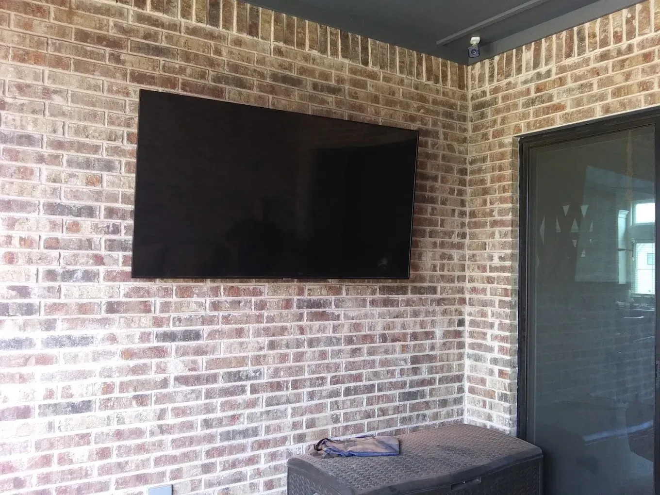 Large TV mounted on a brick wall in a corner of a room, with a dark-colored storage bench below.