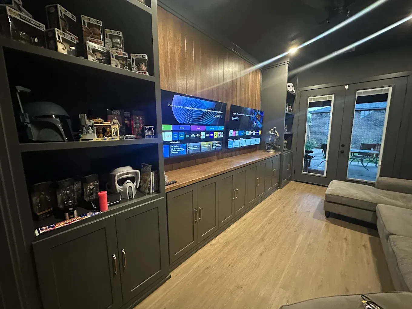 Media room with built-in shelves displaying Star Wars collectibles, three TVs, wooden accents, and a gray sectional sofa.