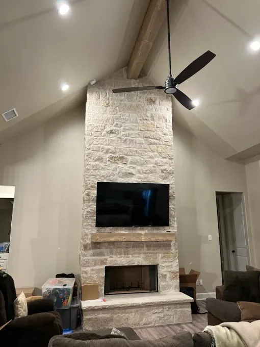 Stone fireplace with mounted TV, fan, and wooden beam in a light-colored room.