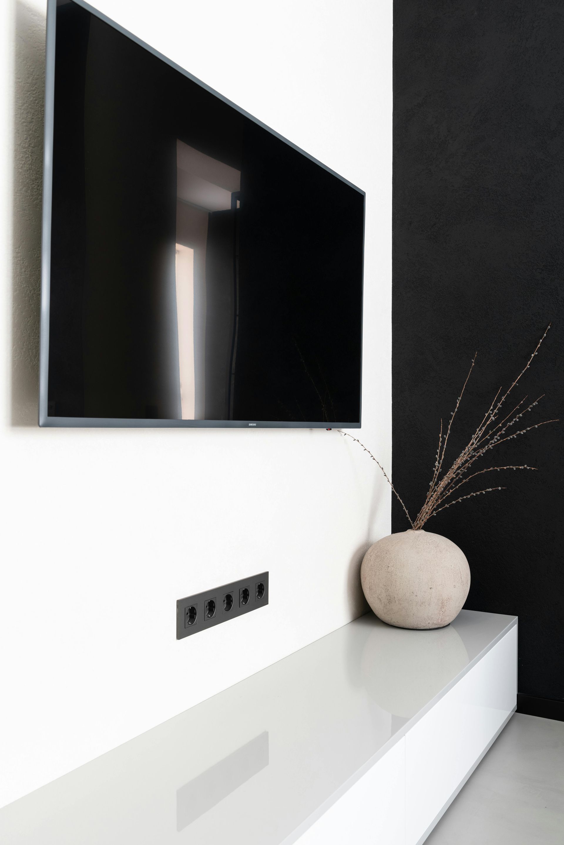 Large TV mounted on a white wall, above a white shelf with a vase and power outlets. Black accent wall.