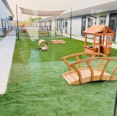 Outdoor daycare play area with artificial turf, wooden climbing toys, and shade sails beside a building