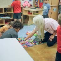 Teacher kneels beside children playing a colorful board game in a classroom