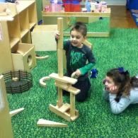 Two children play with a wooden block structure on green carpet in a classroom.