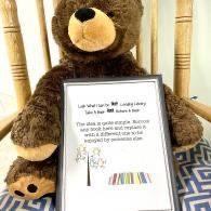 Large brown teddy bear on a chair beside a framed sign with a rainbow stripe and small tree illustration