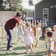 Teacher leading children in an outdoor game on grass near a fenced yard and building