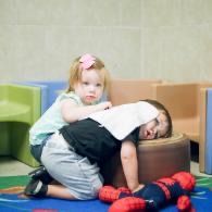 Child in green shirt hugs a child in a white shirt and black pants who is kneeling on a colorful play mat.