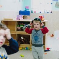 Child indoors holding hands to head, standing in a playroom with shelves and toys in the background