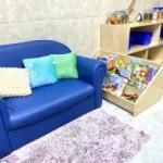 Blue couch with colorful pillows beside a toy shelf and purple rug in a bright playroom