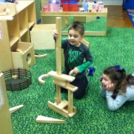 Two children play with wooden blocks on a green carpet in a classroom.