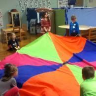 Children in a classroom holding a colorful parachute play circle around it