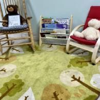 A cozy children’s corner with wicker chairs, a toy shelf, and a green animal-themed rug.