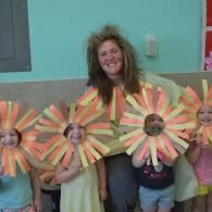 Children posing in flower costumes with smiling center faces in a classroom with a woman behind them