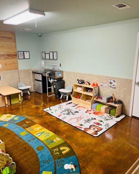 Small classroom with rugs, toy shelves, stools, and a desk against pale green walls.