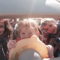 Three children leaning in close inside a car, smiling and holding a yellow ring toy