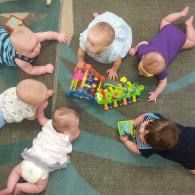 Six babies sitting in a circle, reaching toward a colorful toy on a carpet.