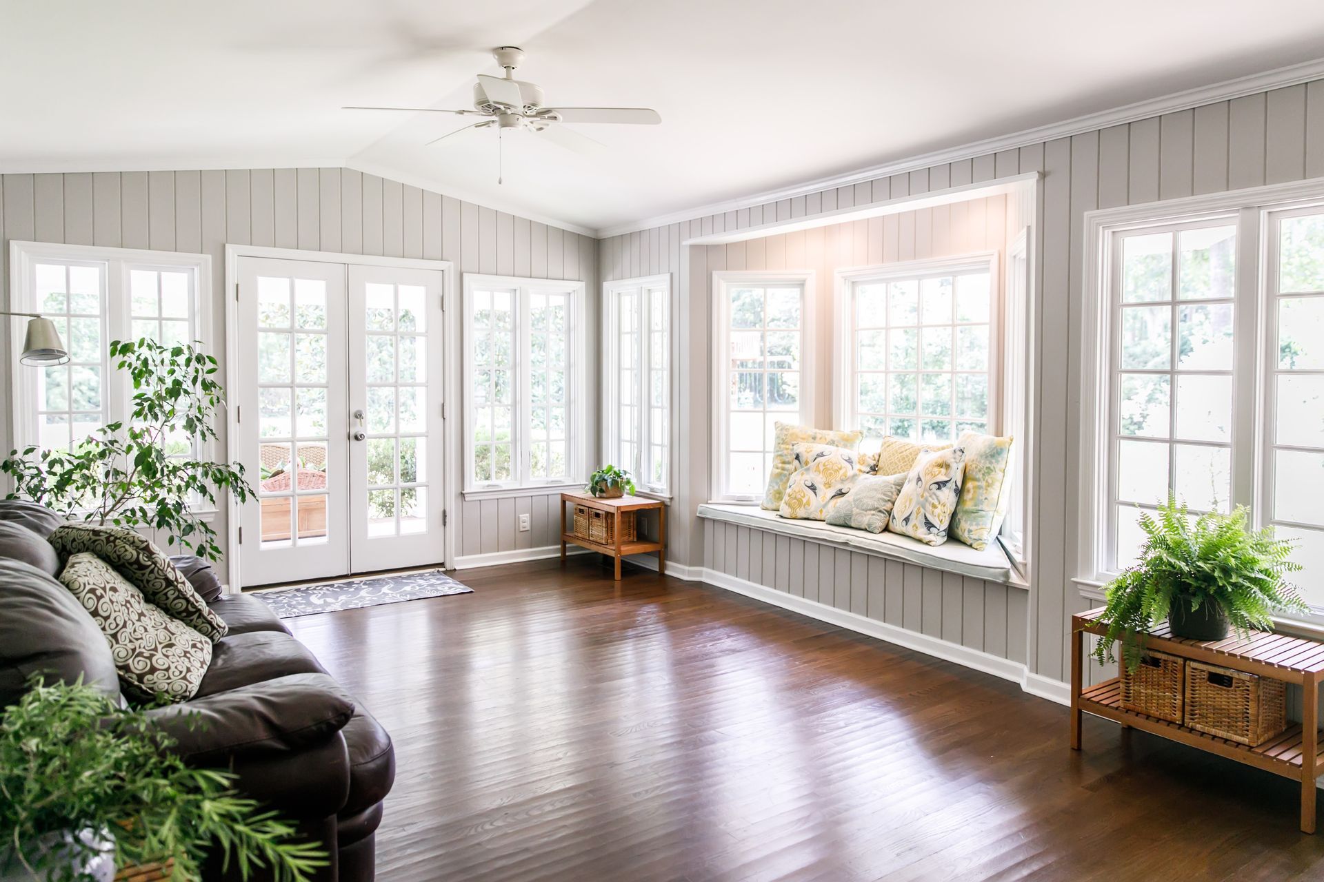 Bright sunroom with dark hardwood floors, white French doors, and a window seat with cushions and plants