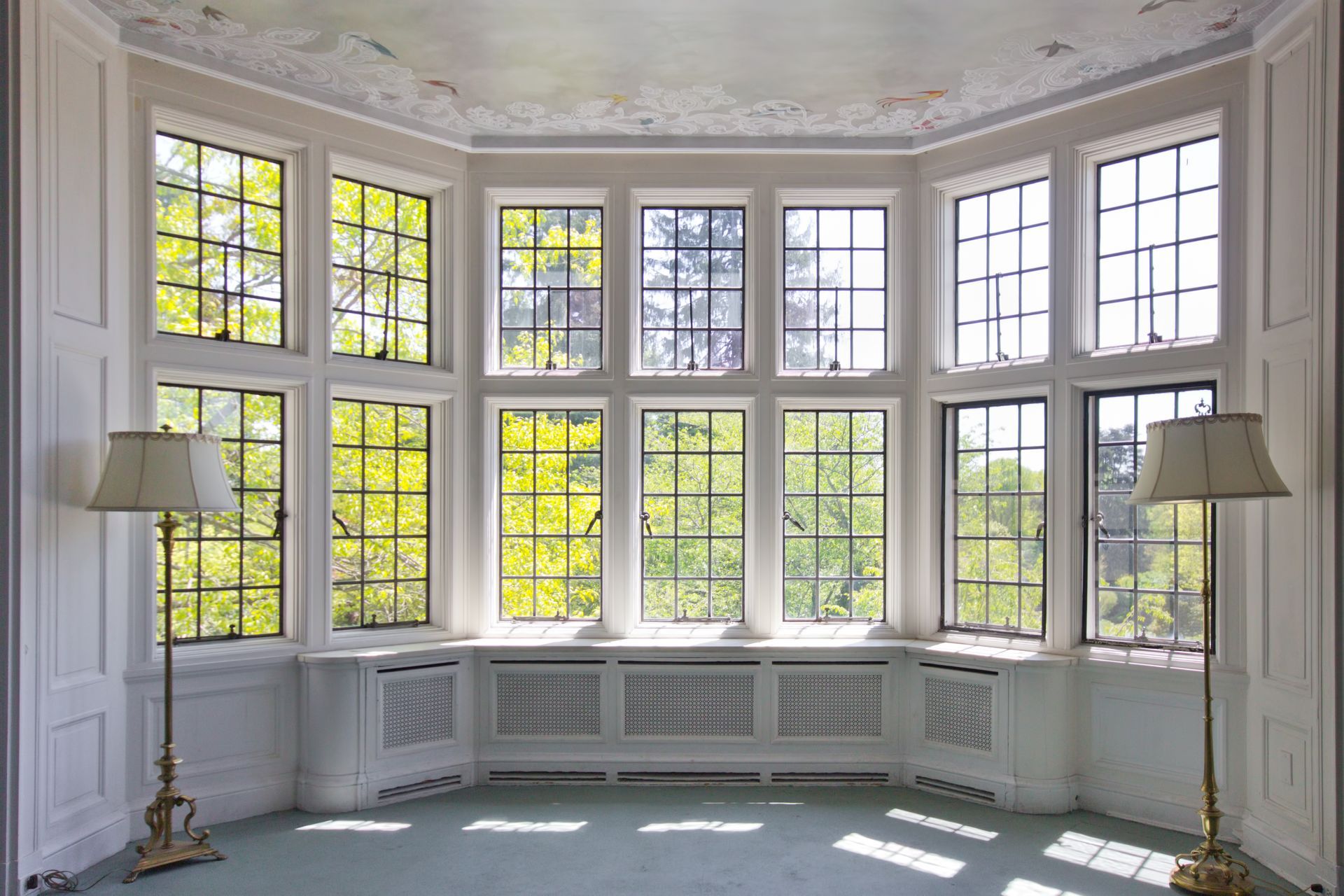 Bright bay-windowed room with white trim, green tree view, and two floor lamps.
