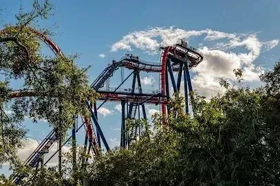 Rollercoaster with blue and red track, high in the sky, behind green trees, against a cloudy sky.