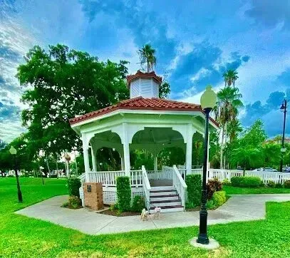 Hvac-Contractor-Bradenton-White gazebo with red roof in a park, surrounded by green grass, trees, and a cloudy blue sky.