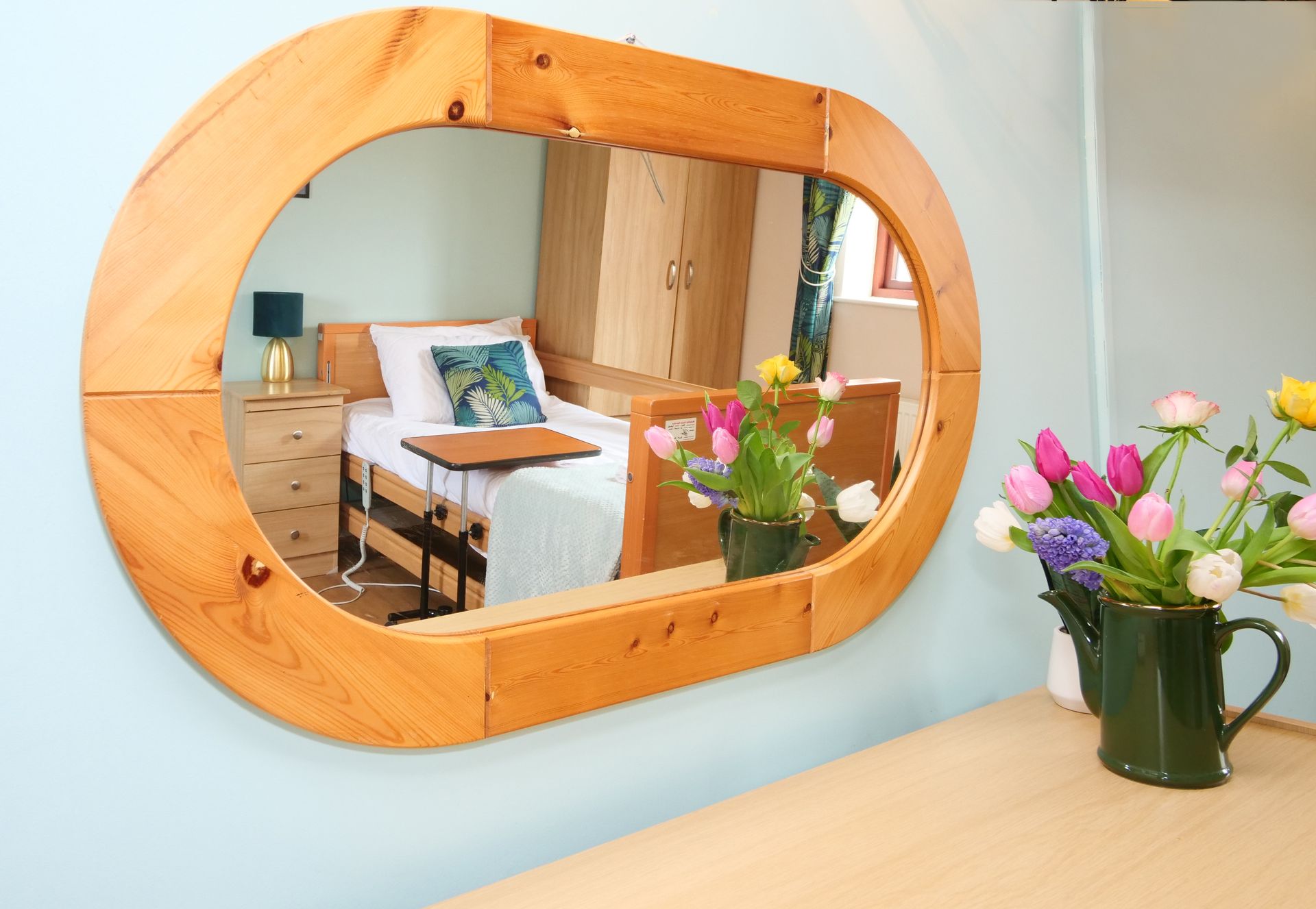A mirror with a wooden frame shows a bedroom with a bed and a table