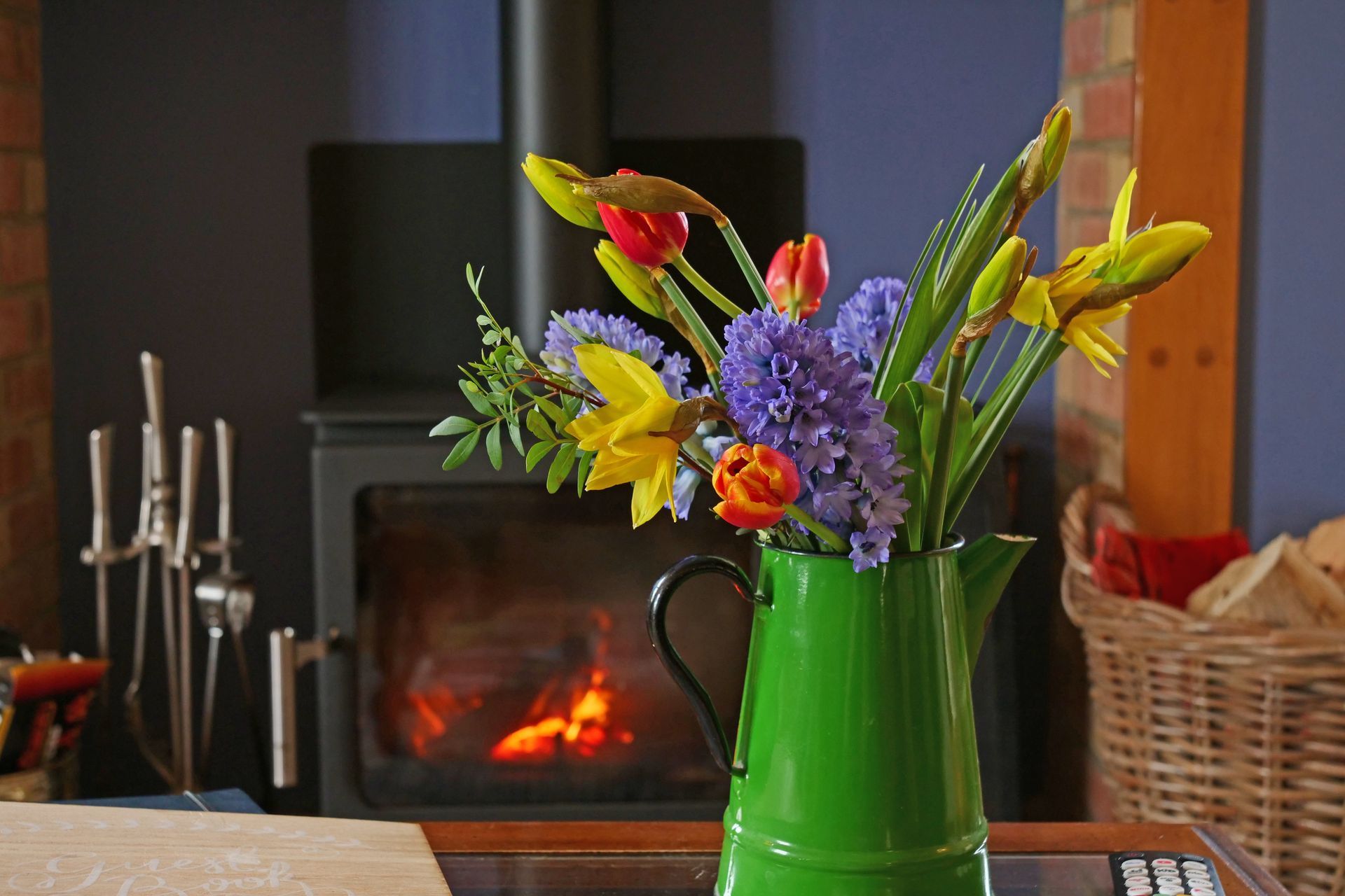 A green pitcher filled with flowers is on a table in front of a fireplace.