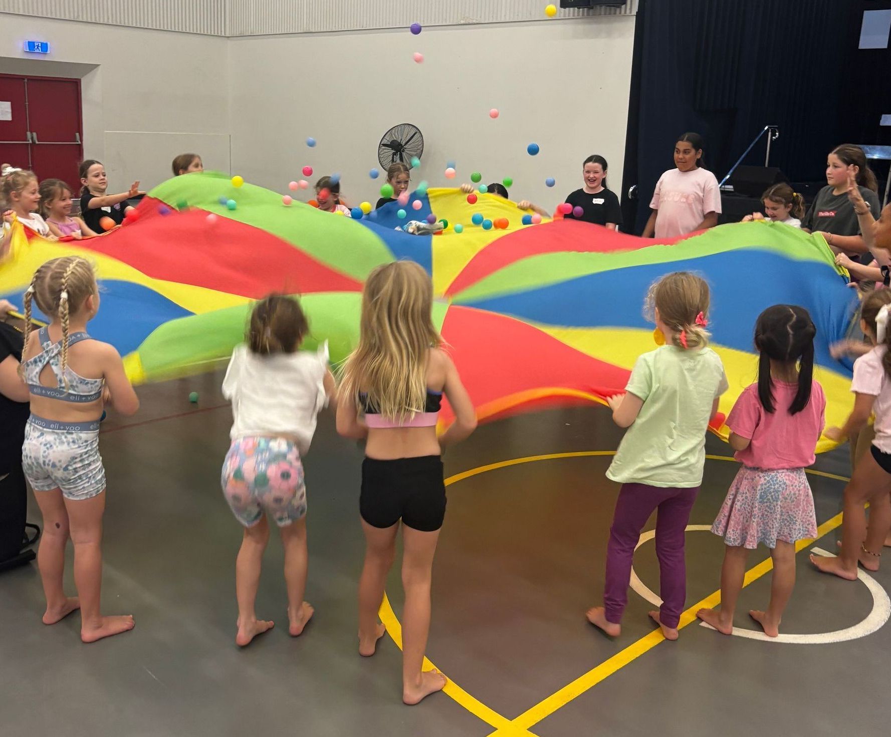 Children playing with a colorful parachute, inside a gymnasium. Balls are tossed into the air.