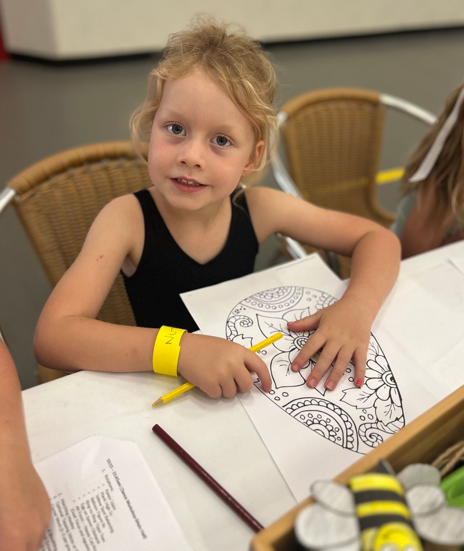 A young girl with blonde curly hair, coloring at a table, smiles at the camera.