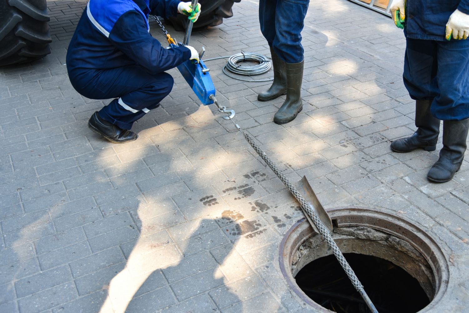 Workers inspecting a manhole, using equipment on a brick-paved surface.