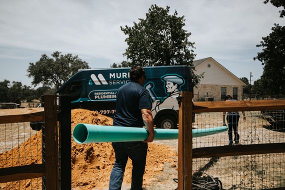 Plumber carrying a long blue pipe, near a truck, fence, and house.