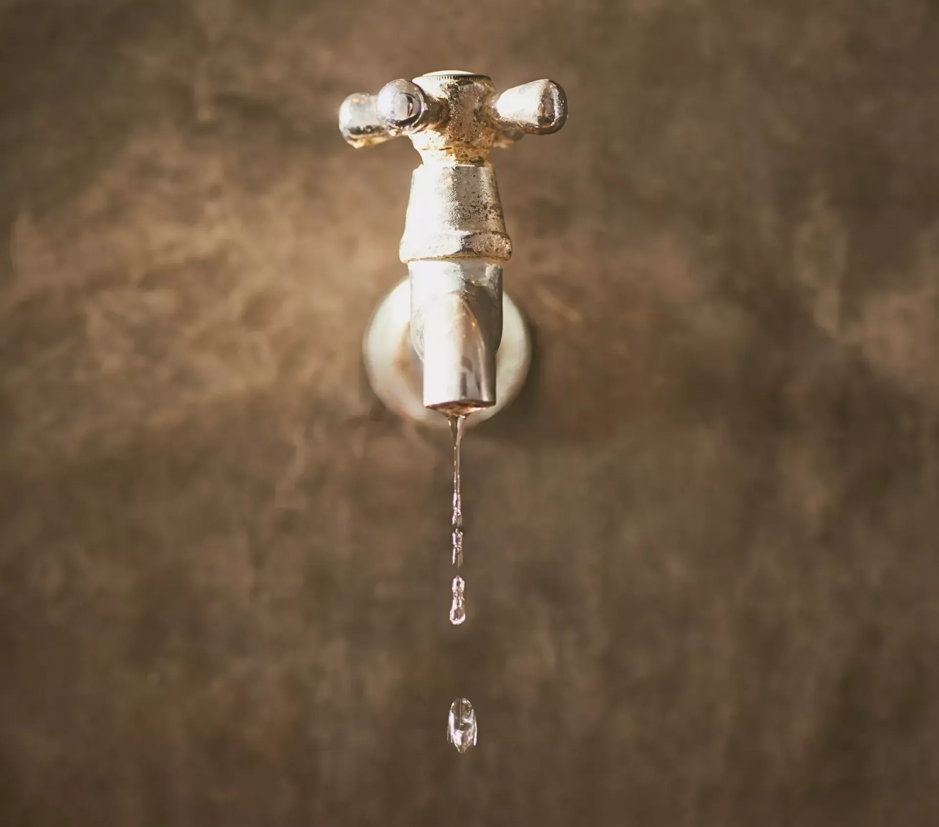 Dripping faucet with water droplets, mounted on a brown wall.