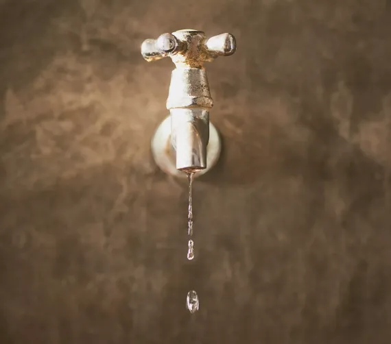 Dripping faucet with water droplets, mounted on a brown wall.