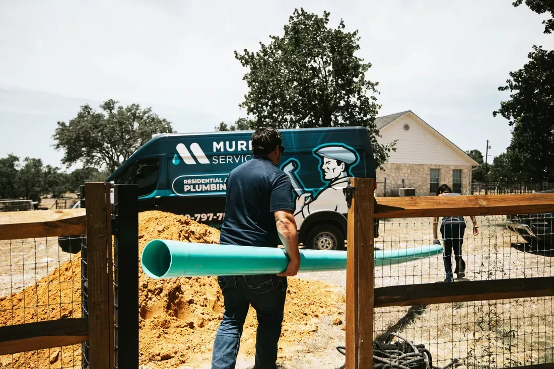Man carrying a large turquoise pipe at a construction site with a plumbing van in the background.