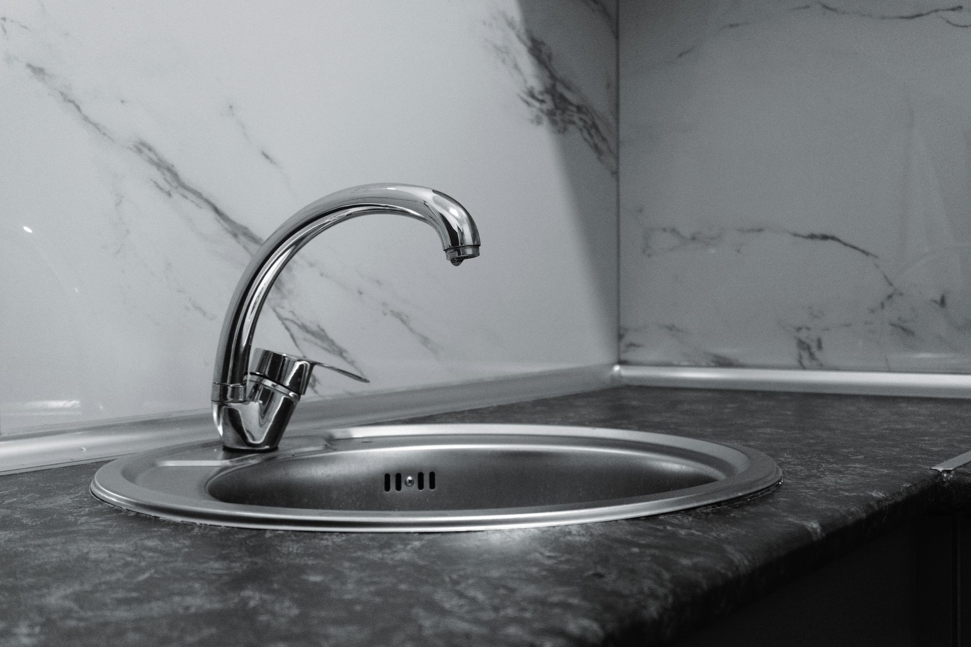 Stainless steel sink and faucet in a kitchen corner with marble-patterned backsplash and dark countertop.