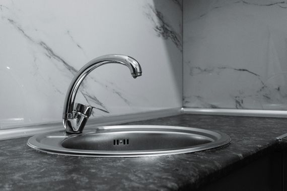 Stainless steel sink and faucet in a kitchen corner with marble-patterned backsplash and dark countertop.