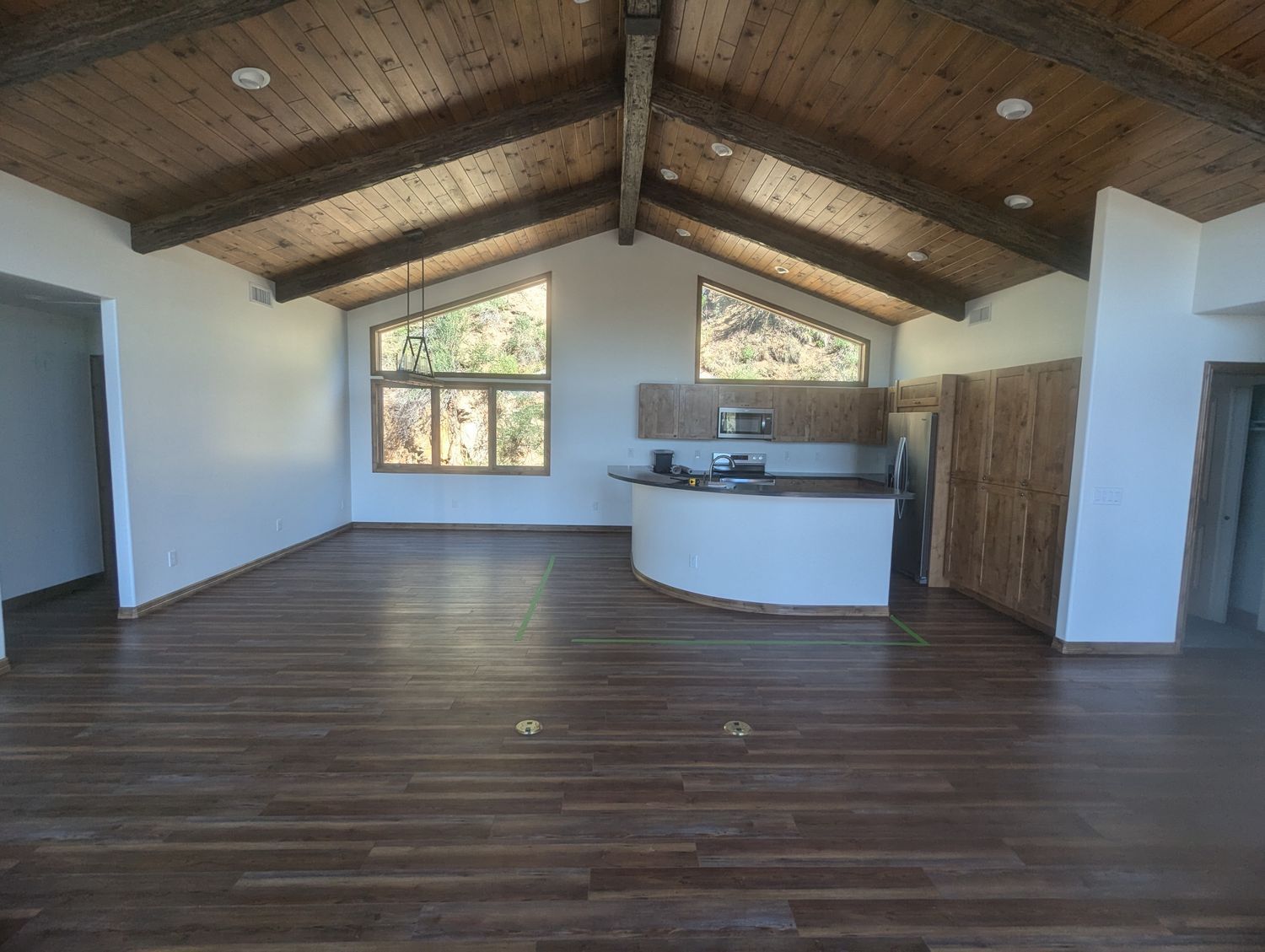 Empty room with wood floors, vaulted wood ceiling, white walls, and a small kitchen.