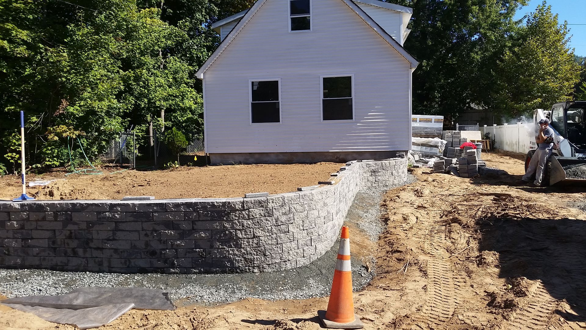A retaining wall made of stacked stone blocks under construction in front of a white house, with a traffic cone nearby.