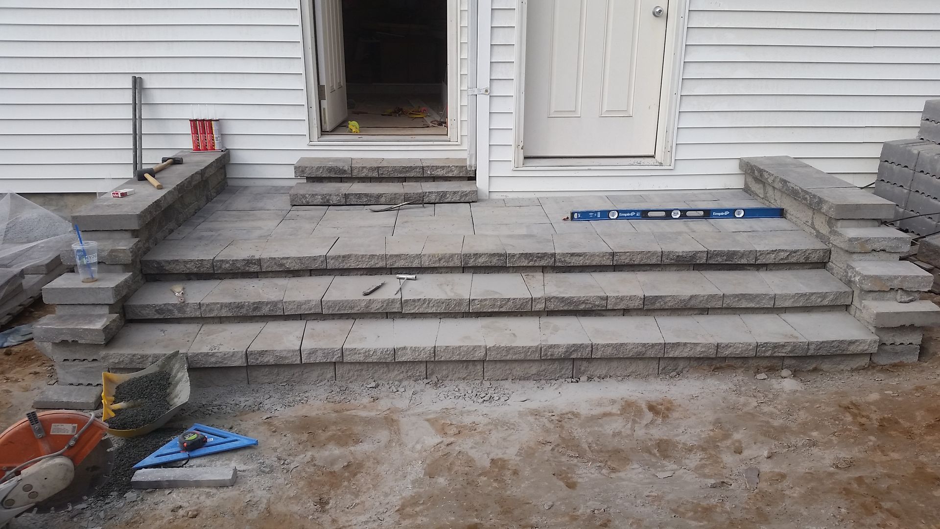 A partially finished grey stone staircase leading to a white-sided house entrance, with construction tools nearby.