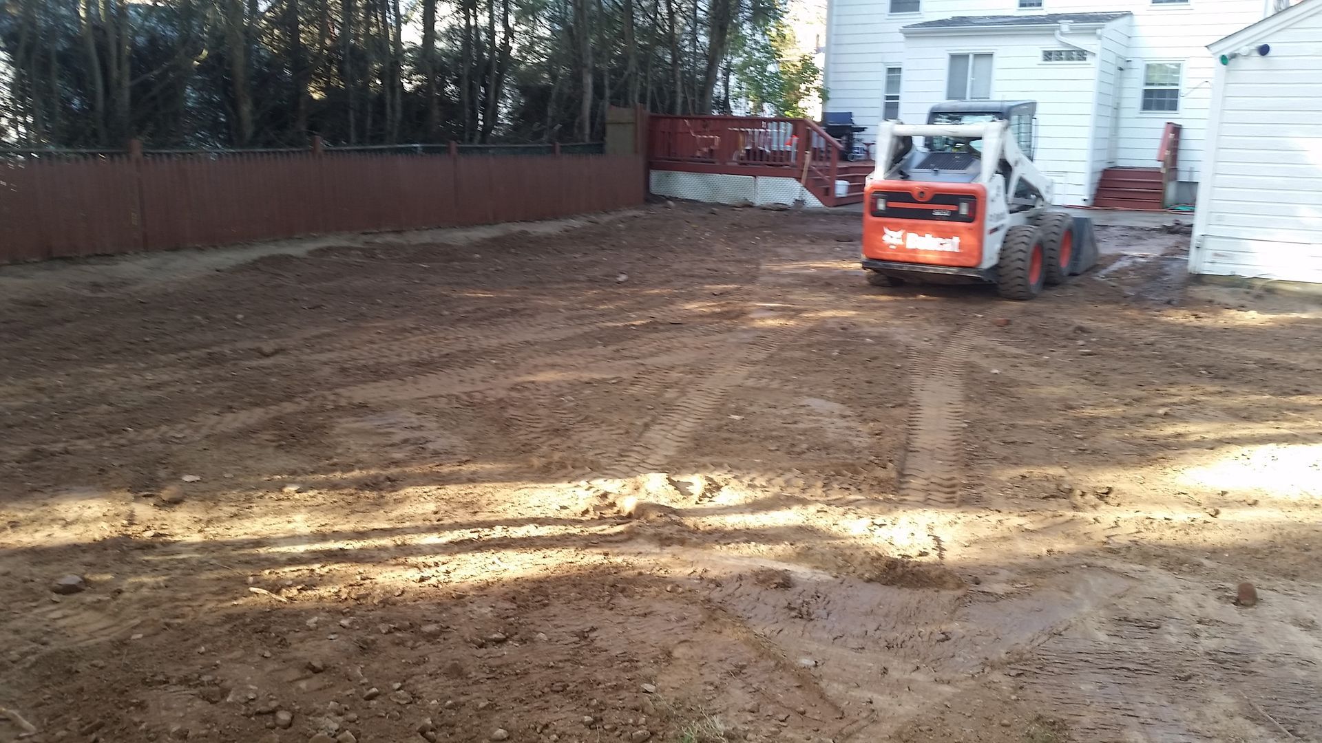 A white Bobcat skid-steer loader sits on a patch of loose, leveled dirt in a backyard near a house and fence.