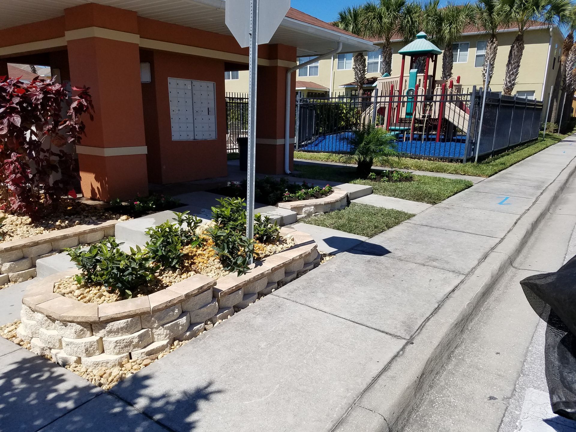 A landscaped planter with small shrubs sits on a sidewalk near a community mail station and a playground.